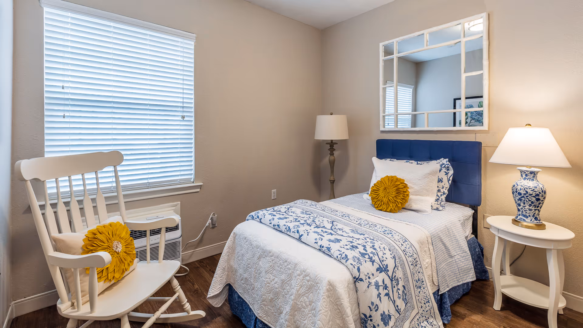 A cozy bedroom with a single bed featuring blue and white floral bedding and a yellow decorative pillow. Next to the bed is a white round side table with a blue and white patterned lamp. A tall floor lamp stands in the corner. A white rocking chair with a yellow flower-shaped pillow is placed near a window with white blinds. A large decorative mirror hangs on the wall above the bed.