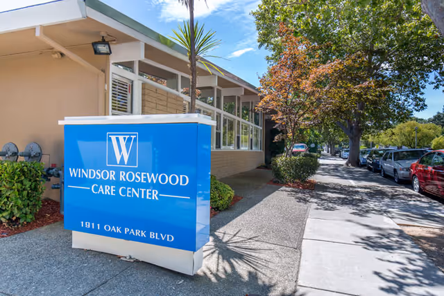 Exterior view of Windsor Rosewood Care Center showing a blue sign with the facility name and address 1911 Oak Park Blvd, a sidewalk, parked cars, and trees lining the street on a sunny day.