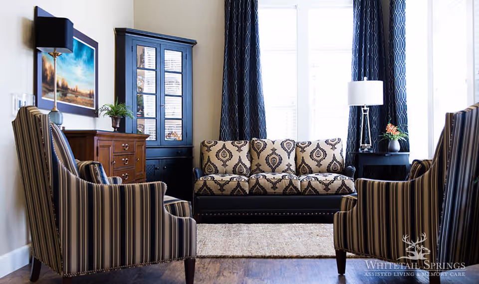 A cozy living room with a patterned beige and black sofa centered between two large windows with dark blue curtains. Two striped armchairs face the sofa, and there is a wooden cabinet with a plant and a lamp on the left side. A small black side table with a lamp and a flower vase is on the right side. The floor is wooden with a light-colored rug in the middle.