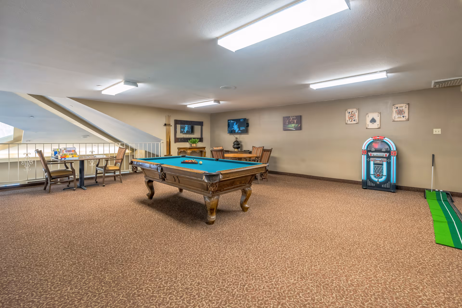 A spacious game room with a pool table in the center, a small table with board games and chairs near a railing, a wall-mounted TV, framed pictures on the wall, a jukebox, and a putting green on the right side.