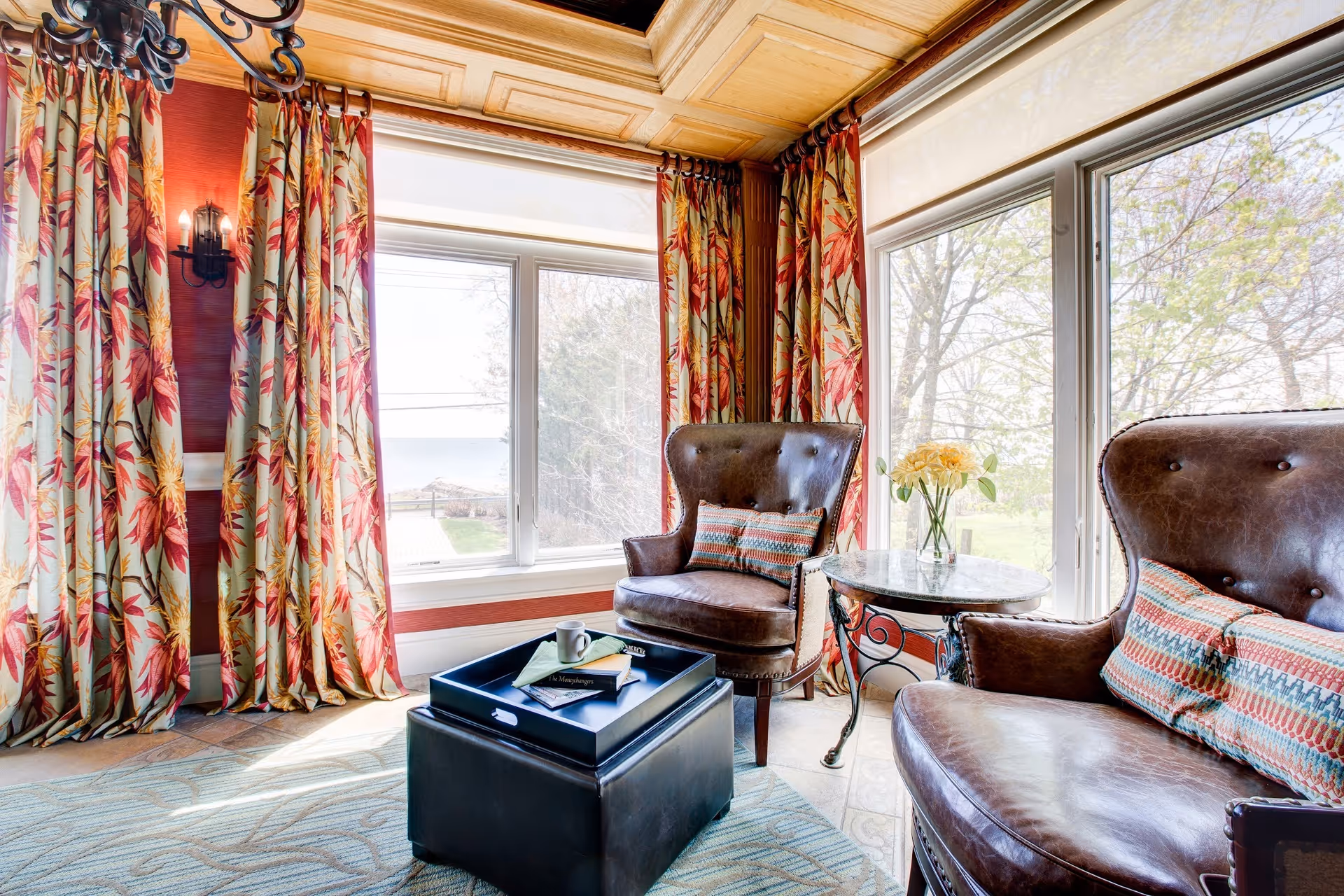 Sunlit sitting area with two leather armchairs, a small round table with flowers, an ottoman with a tray, and large windows with patterned curtains.