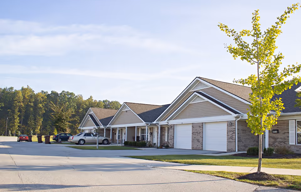 Exterior view of a senior living facility with multiple attached units featuring garages, driveways, and a young tree in the foreground. Several cars are parked along the driveway, and there are trees in the background under a partly cloudy sky.