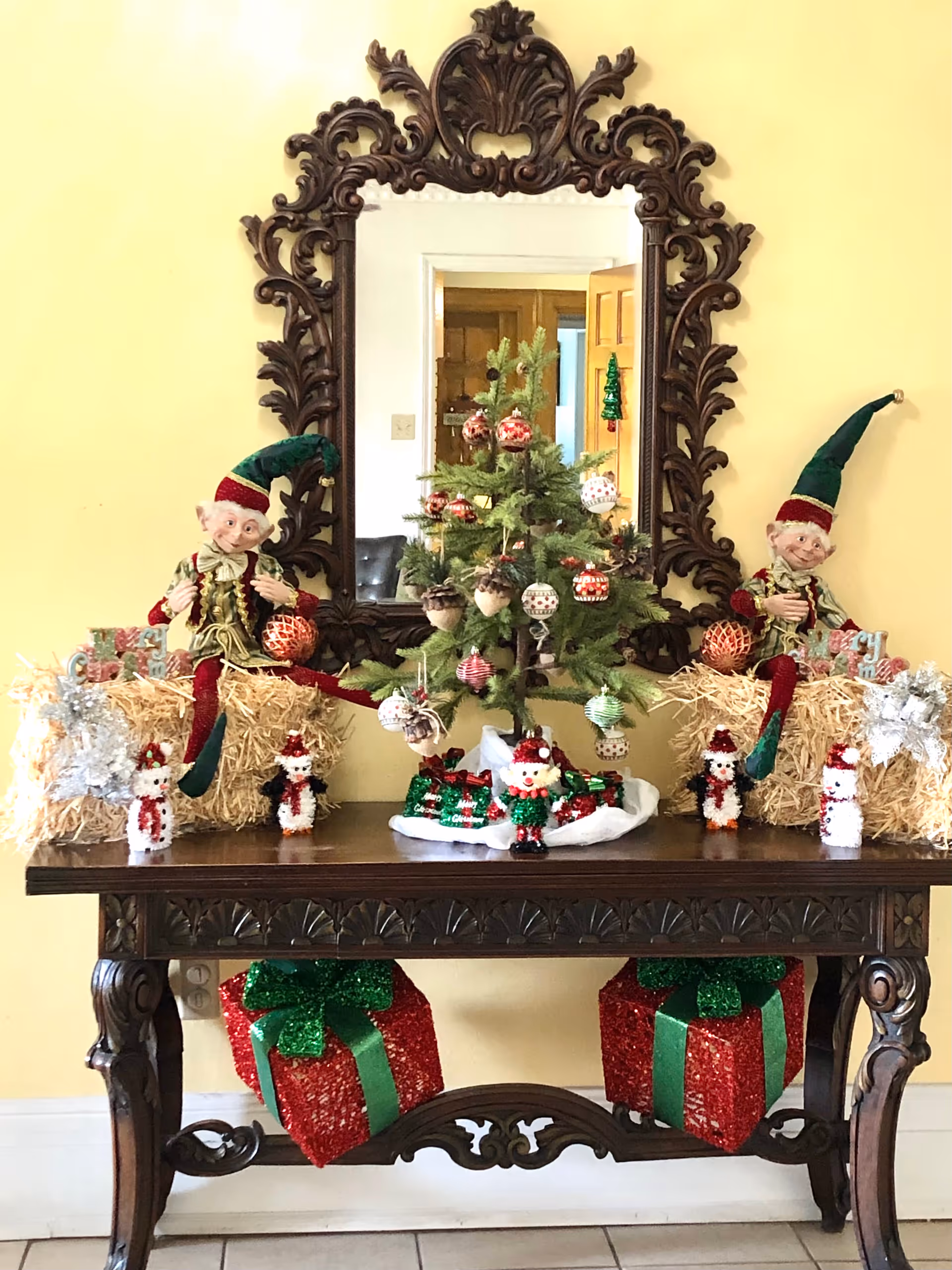 A decorative wooden table with Christmas decorations including a small decorated Christmas tree, two elf dolls sitting on hay bales, small snowman figurines, and wrapped gift boxes underneath. A large ornate mirror hangs on the yellow wall behind the table, reflecting part of the room.
