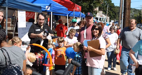 A group of people gathered outdoors at a community event with tents and booths. Several children and adults are interacting, with one man holding balloon animals. The atmosphere appears lively and social.