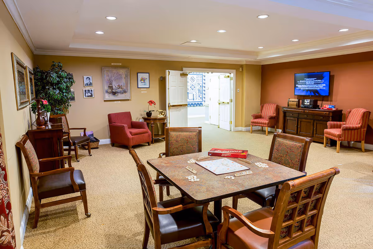 Well-lit common room with a central game table and chairs, armchairs and a TV against the wall, and open double doors in the background.