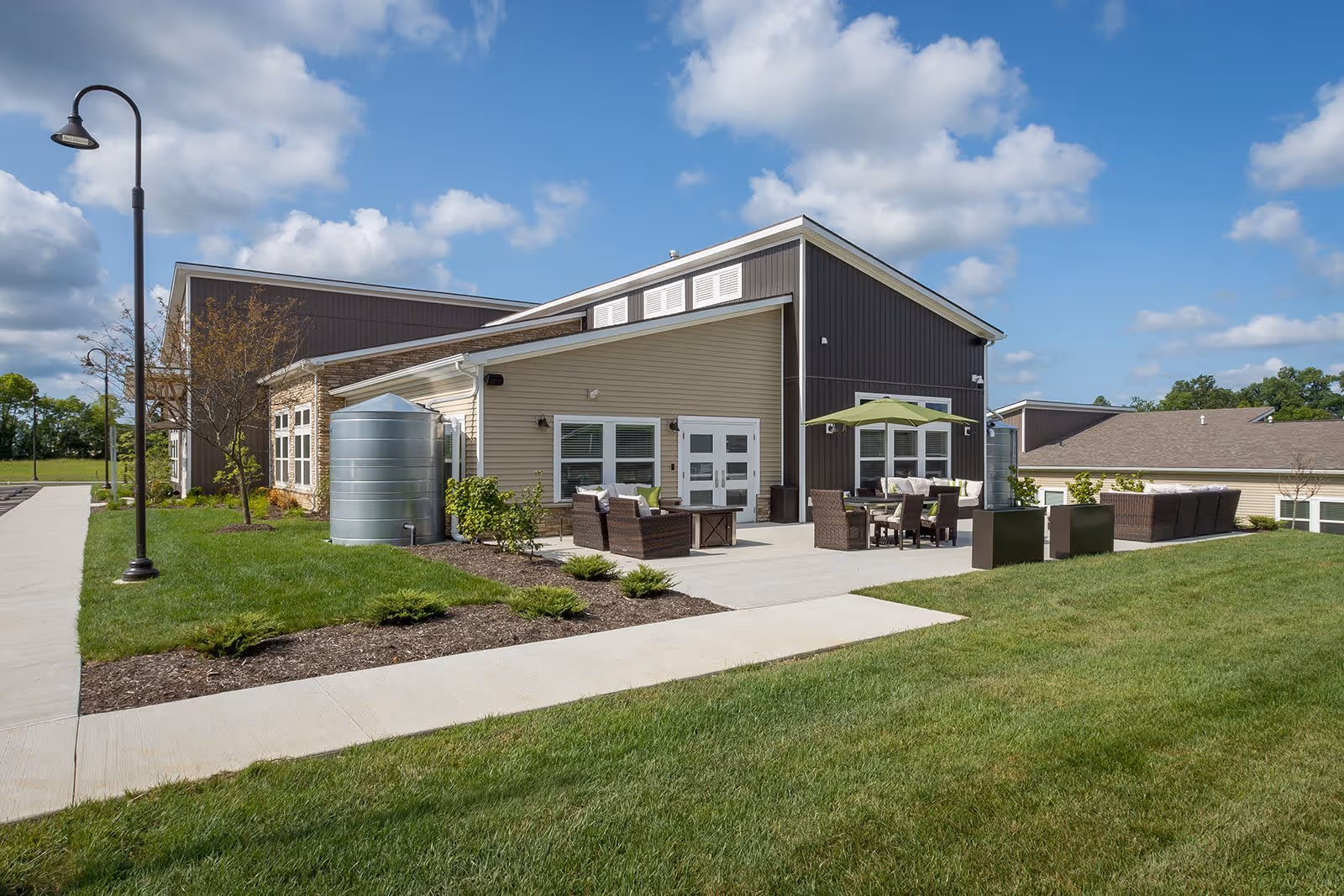 Outdoor patio area of a senior living facility with wicker furniture including chairs and sofas, a table with an umbrella, and well-maintained landscaping. The building has beige and dark brown siding with white trim, and the sky is partly cloudy.