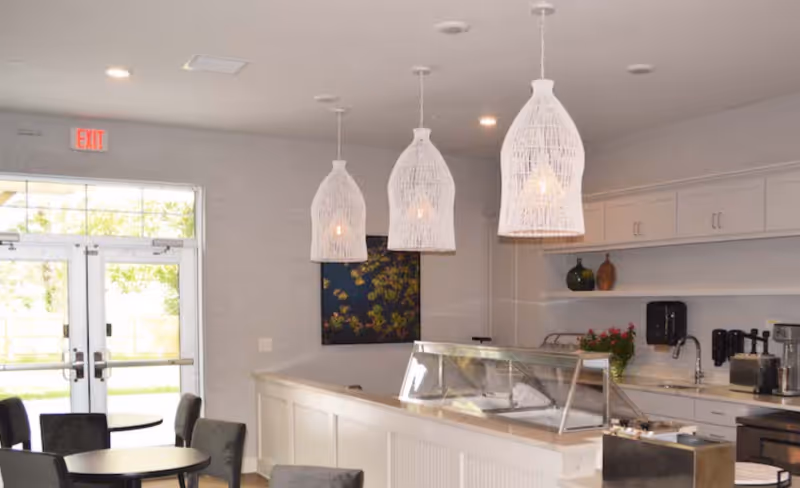 Bright communal dining and serving area with round tables, a service counter with glass display, white pendant lights, and double glass doors.