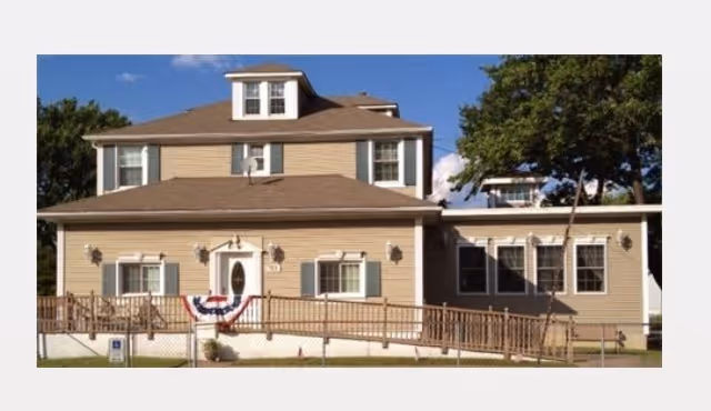 Front exterior of a beige two-story assisted living building with a wheelchair ramp, porch, and patriotic bunting.