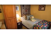 A cozy bedroom with a single bed featuring a colorful patterned blanket and several pillows, including a teddy bear. There is a wooden wardrobe on the left side and a nightstand with a lamp next to the bed. The walls are painted yellow and decorated with framed pictures.