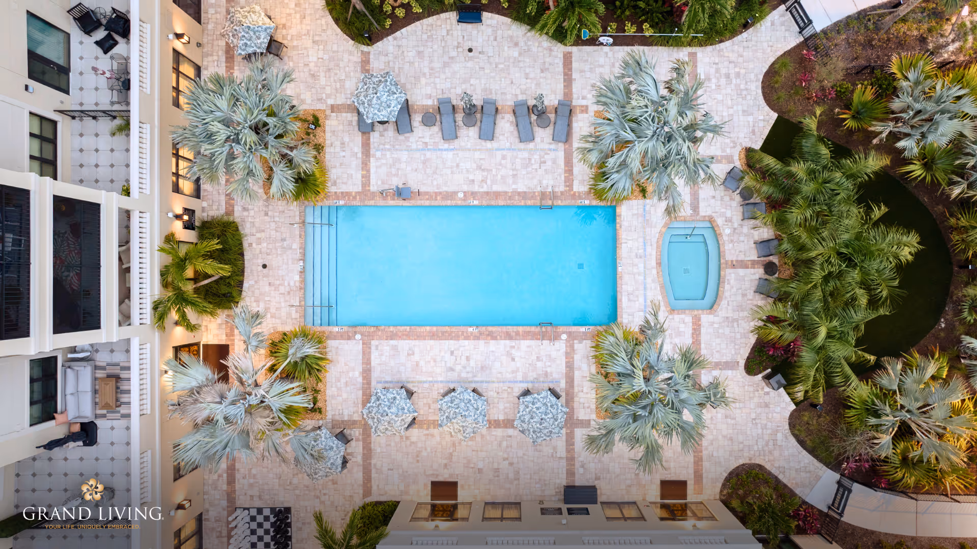 Aerial view of a resort-style courtyard with a rectangular swimming pool, adjacent hot tub, lounge chairs, umbrellas, and palm trees.