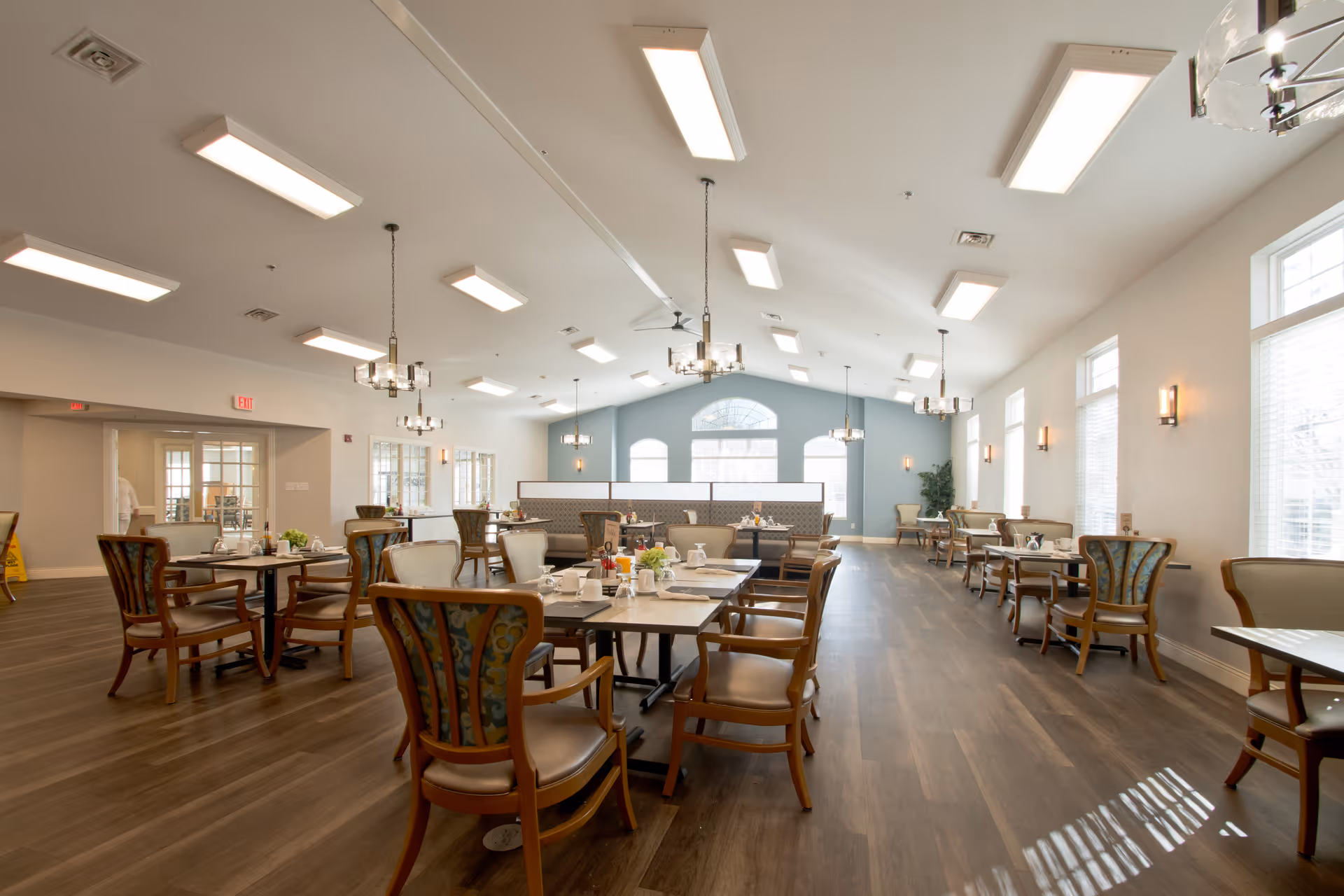 A spacious and well-lit dining room with multiple tables and chairs arranged neatly. The room features large windows allowing natural light to fill the space, modern chandeliers hanging from the ceiling, and a light blue accent wall at the far end. The tables are set with plates, cups, and small flower arrangements, creating an inviting atmosphere.