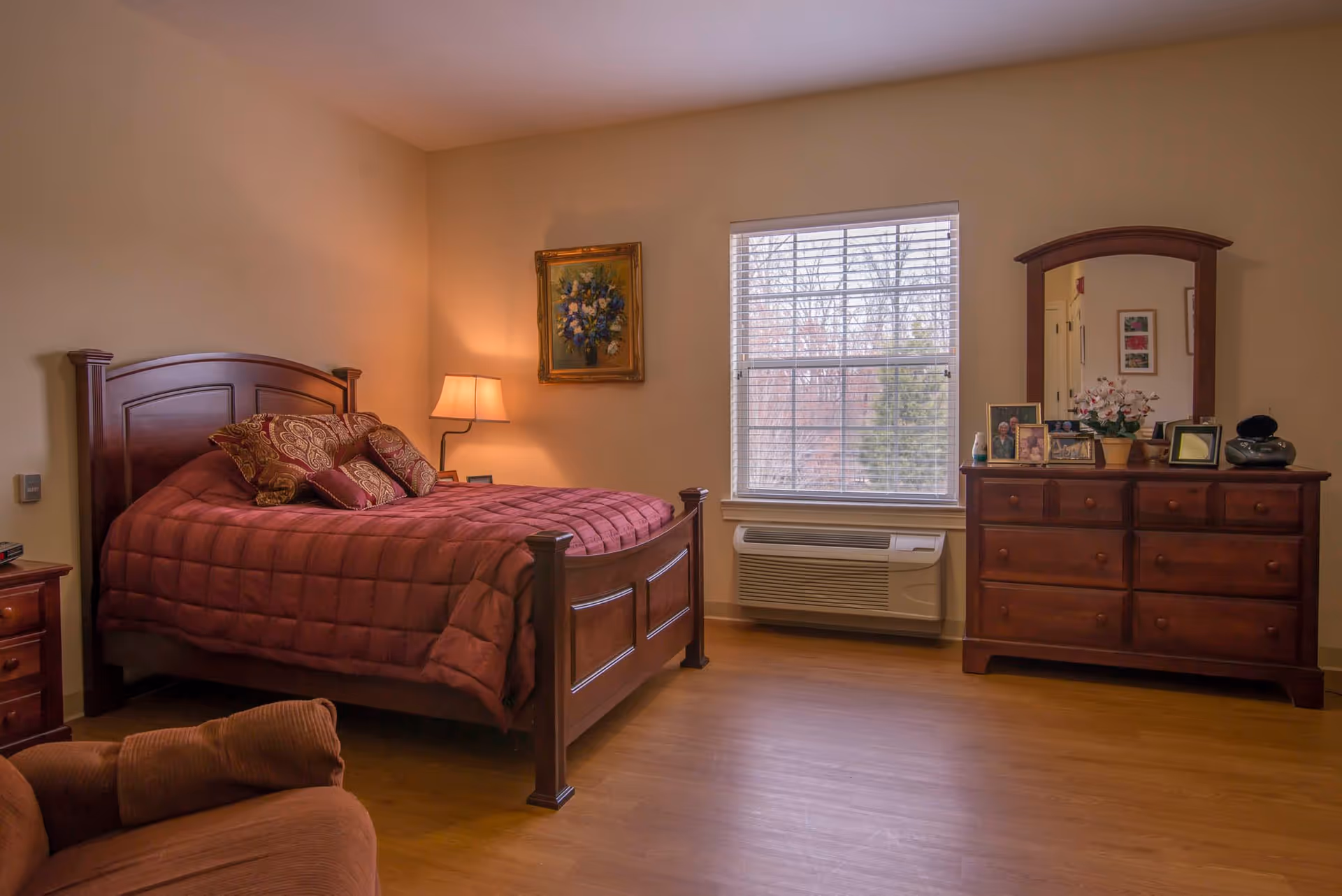 Well-lit bedroom with a wooden bed, nightstand, dresser with mirror, and an armchair.