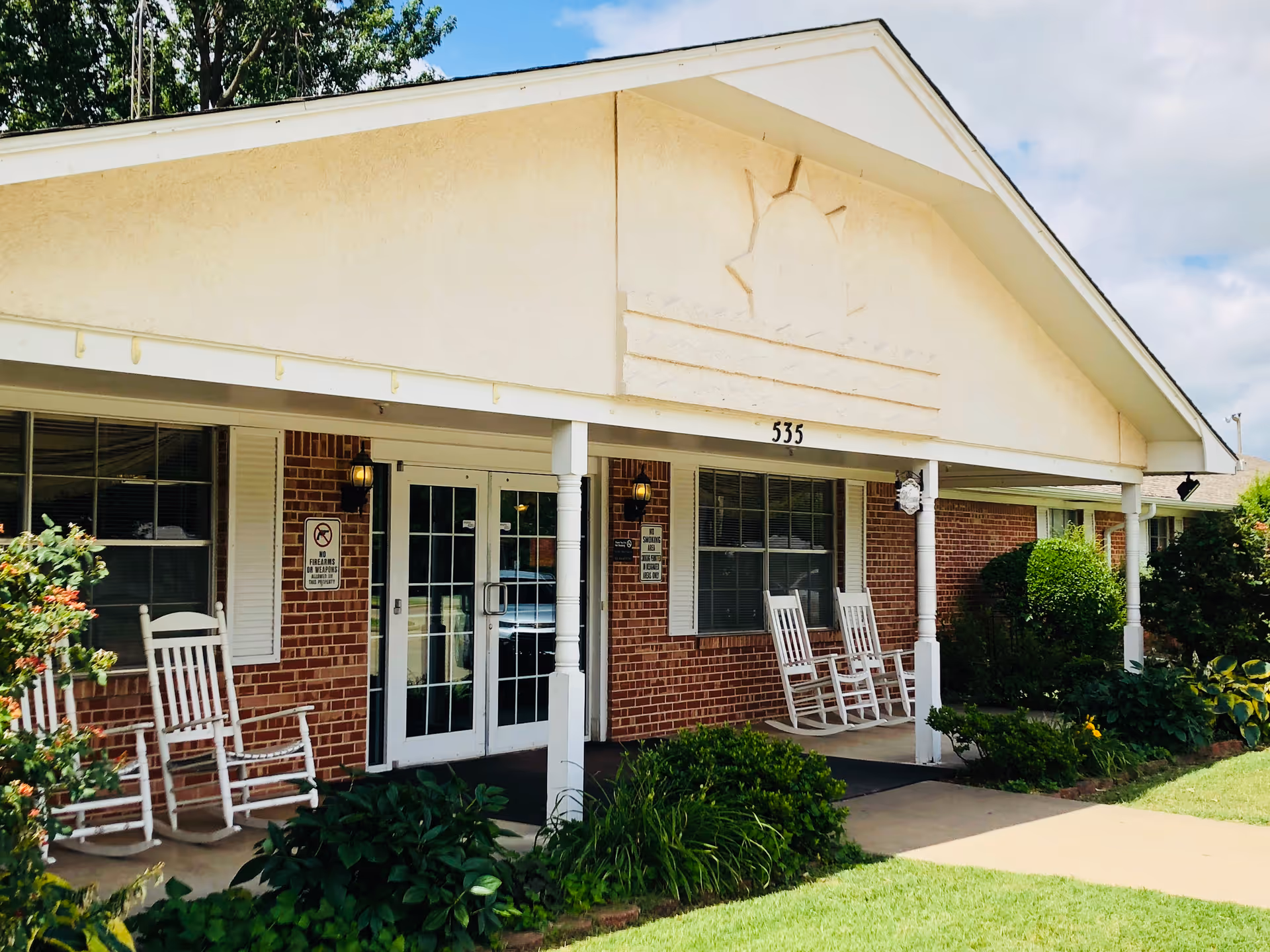 Front exterior view of a single-story brick building with a white porch roof and four white rocking chairs on the porch. The building has large windows with white shutters and a glass double door entrance. The address number 535 is displayed above the entrance. There are green bushes and plants in front of the porch and a well-maintained lawn. The sky is partly cloudy.