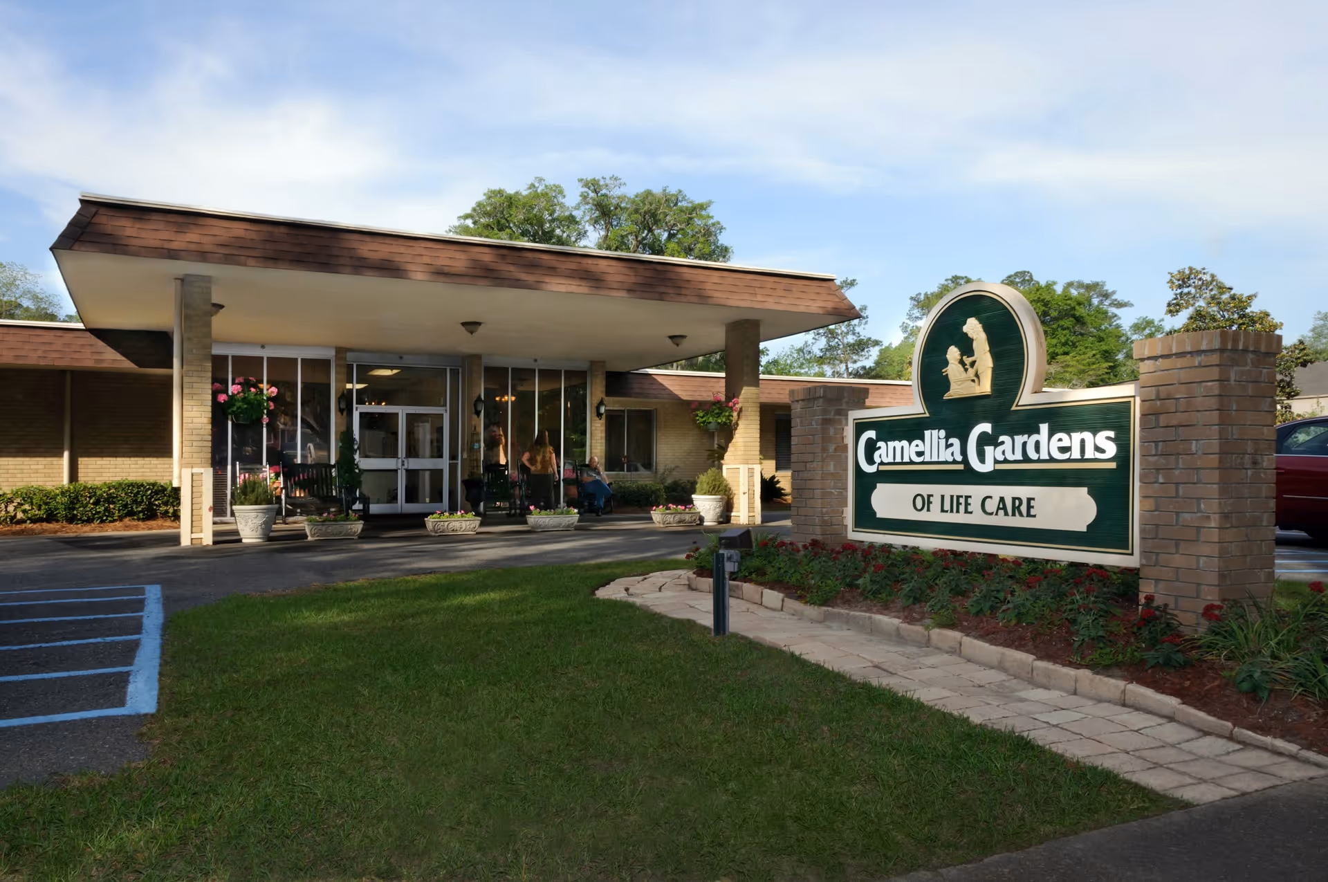 Front entrance of Camellia Gardens of Life Care with a large facility sign, covered porte-cochère, and potted plants.