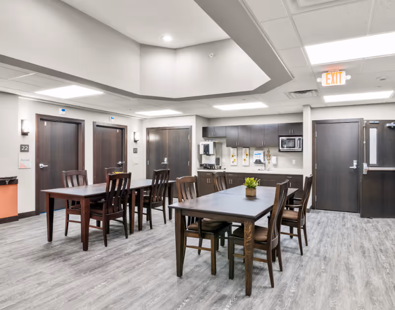 Interior view of a senior living facility common area with two wooden tables and chairs, a small plant on one table, wood-finished cabinets, a coffee machine, microwave, and a sink along the back wall. There are multiple closed doors, light gray walls, and a gray wood-patterned floor. An exit sign is visible on the ceiling near a door.