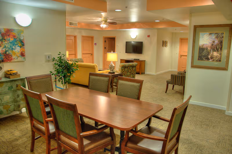Interior view of a senior living facility common area at Lakeside Park featuring a wooden dining table with six chairs in the foreground, a small green chest with decorative items and a plant on the left, and a living room area with a yellow sofa, patterned armchairs, a table lamp, and a wall-mounted TV in the background. The walls are decorated with colorful paintings and the space is warmly lit.