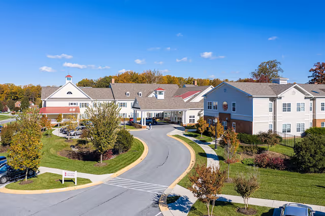 Exterior view of Brooke Grove Rehabilitation and Nursing Center showing a large, multi-wing building with a curved driveway entrance, surrounded by green lawns, trees, and a clear blue sky.