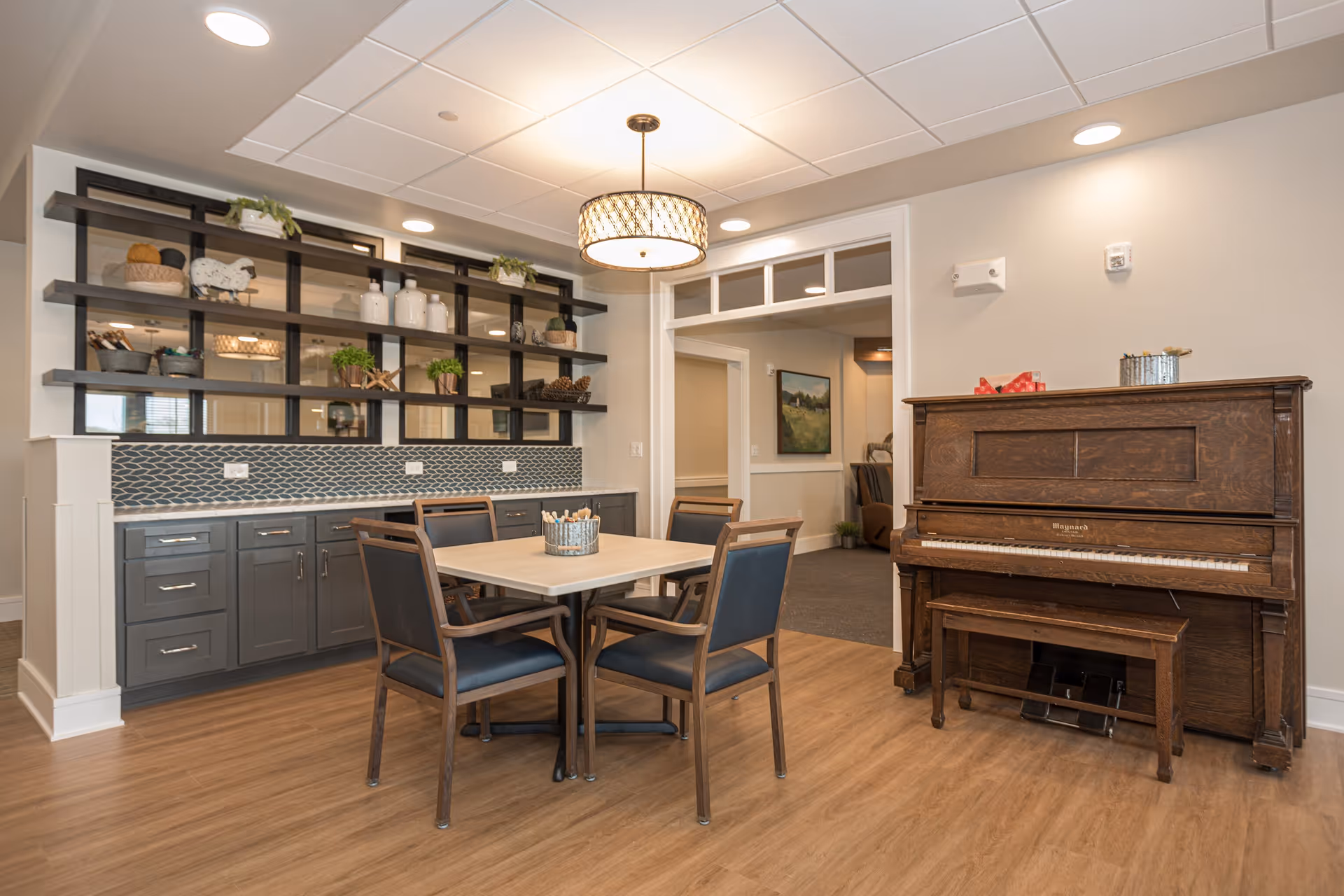 A cozy interior room with a wooden upright piano on the right and a square table with four chairs in the center. Behind the table is a counter with gray cabinets and a decorative backsplash, topped with shelves holding various decorative items and plants. The room has wood flooring, a ceiling light fixture, and an open doorway leading to another room with artwork on the wall.
