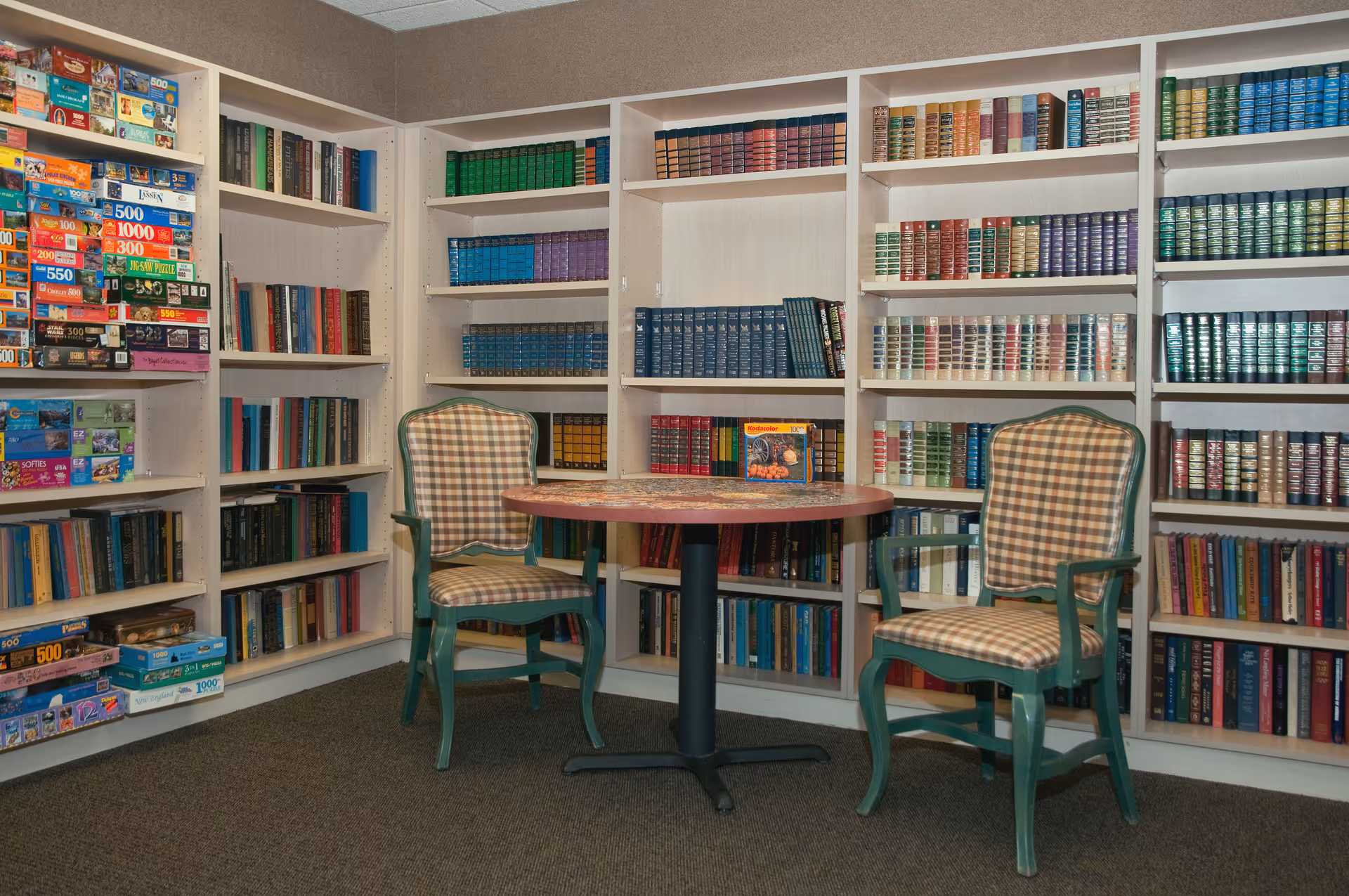 A cozy reading and puzzle area with two green wooden chairs with plaid cushions around a round table. Surrounding the seating area are white bookshelves filled with books and various jigsaw puzzles.