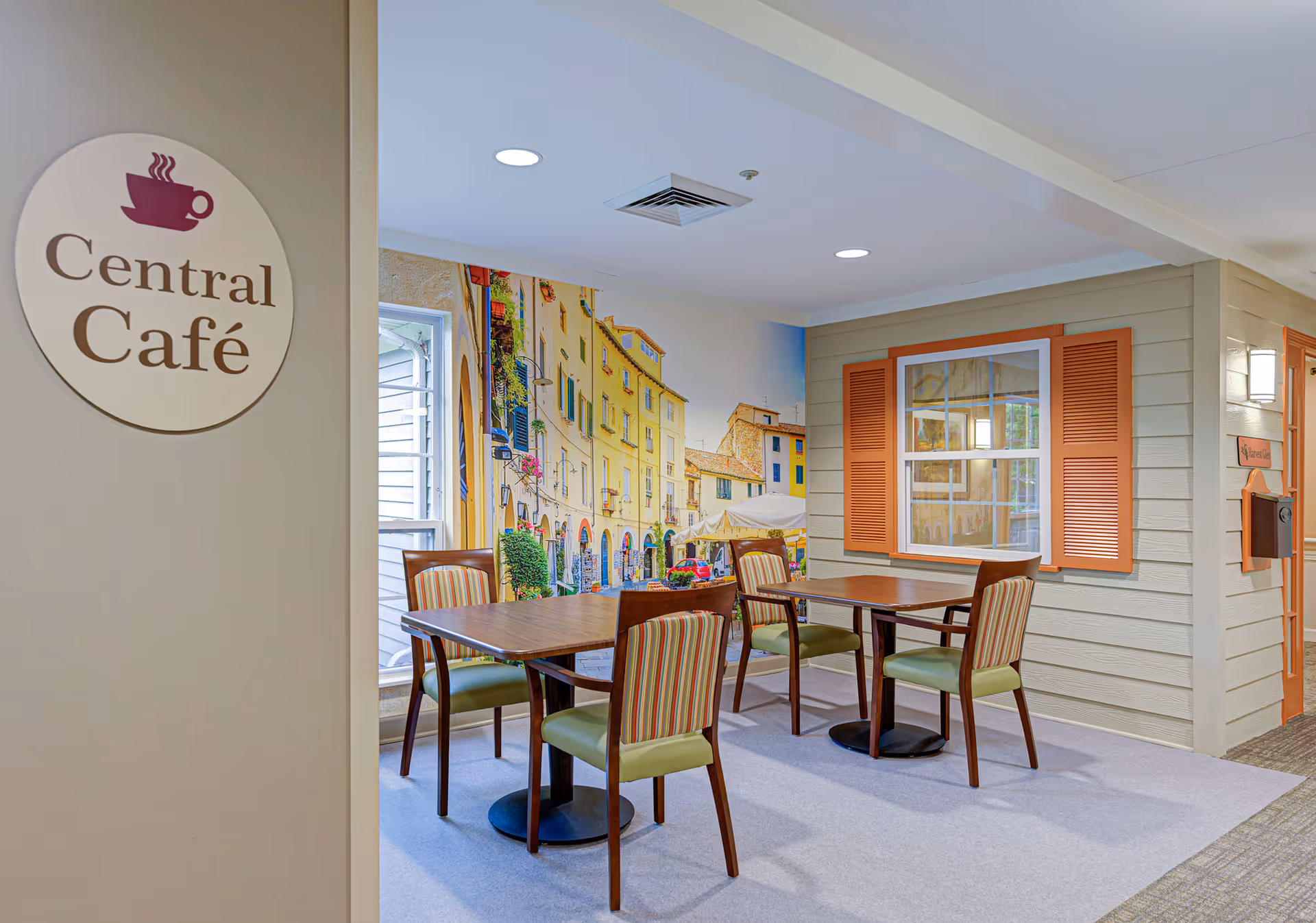 Interior view of a dining area named Central Café with two wooden tables and four chairs with striped upholstery. The walls feature a mural of a European street scene and a decorative window with orange shutters. The space is well-lit with ceiling lights.