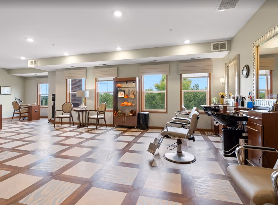 A spacious and well-lit salon area with large windows showing green trees outside. The room features salon chairs with footrests, a hair washing station, a wooden shelving unit decorated with autumn-themed items, and a small seating area with a round table and chairs. The floor has a patterned design, and the walls are painted in a light neutral color.