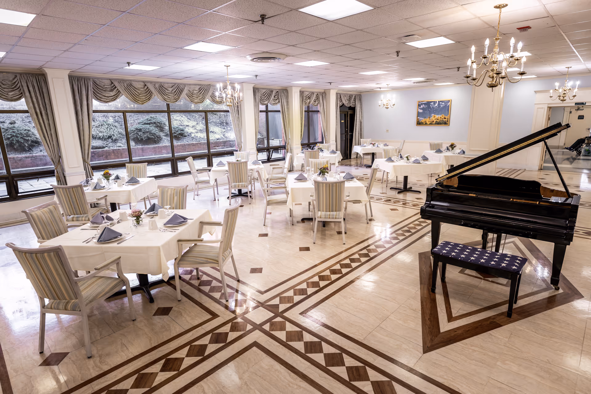 A spacious dining room with multiple tables covered in white tablecloths, each set with napkins and small floral centerpieces. The room features large windows with draped curtains, chandeliers hanging from the ceiling, and a grand piano with a bench in the foreground. The floor has a decorative tile pattern.