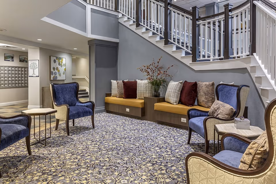 A cozy seating area in a senior living facility featuring blue upholstered armchairs with wooden frames, a built-in bench with mustard yellow cushions and various decorative pillows, a small side table, and a staircase with white railings. The floor is covered with a patterned carpet, and there are framed artworks on the walls near a mail area.