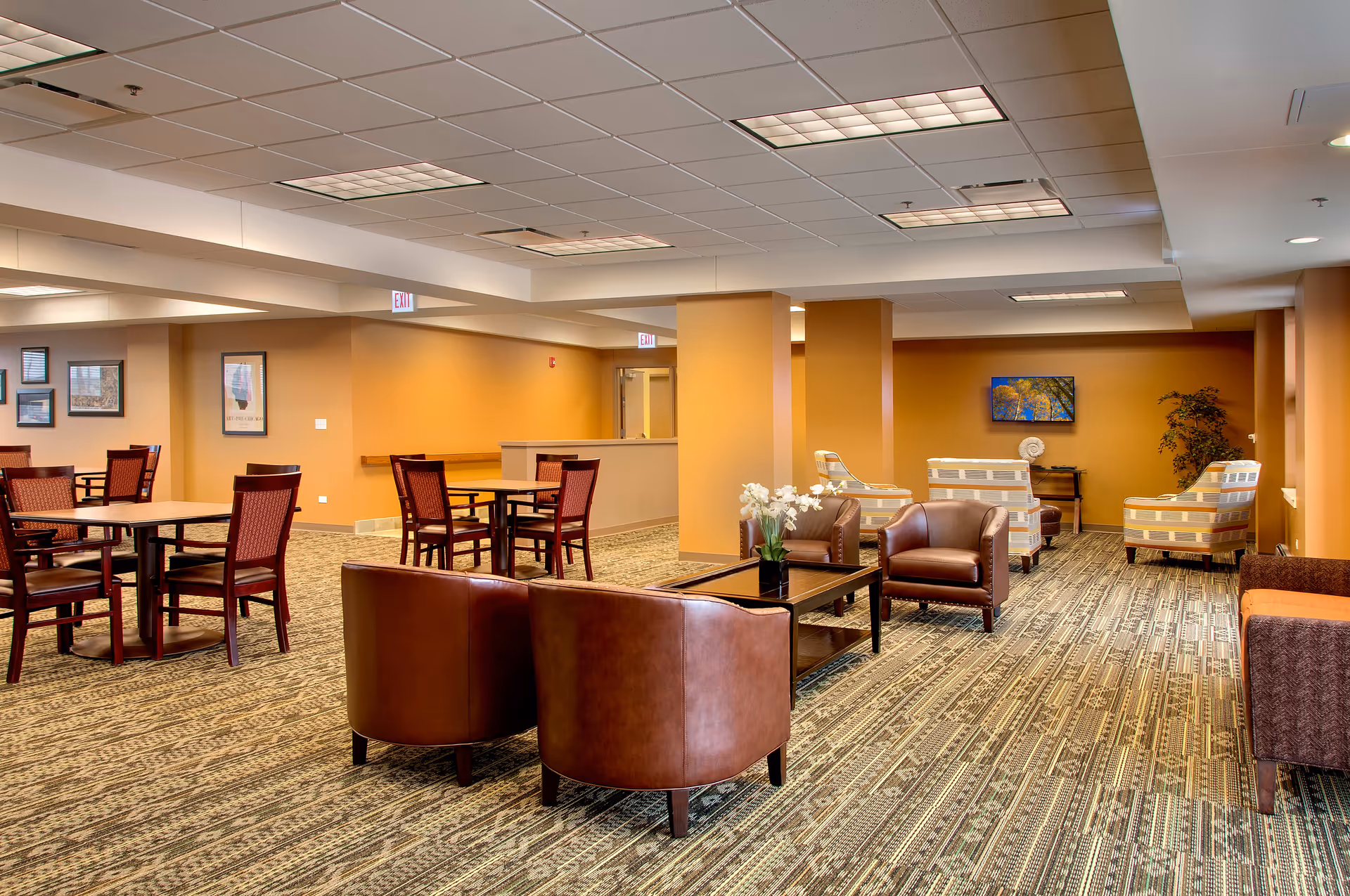 A spacious senior living facility common area with multiple seating arrangements including brown leather chairs, patterned armchairs, and wooden tables with chairs. The room has warm beige walls, patterned carpet, framed artwork on the walls, and ceiling lights providing bright illumination.