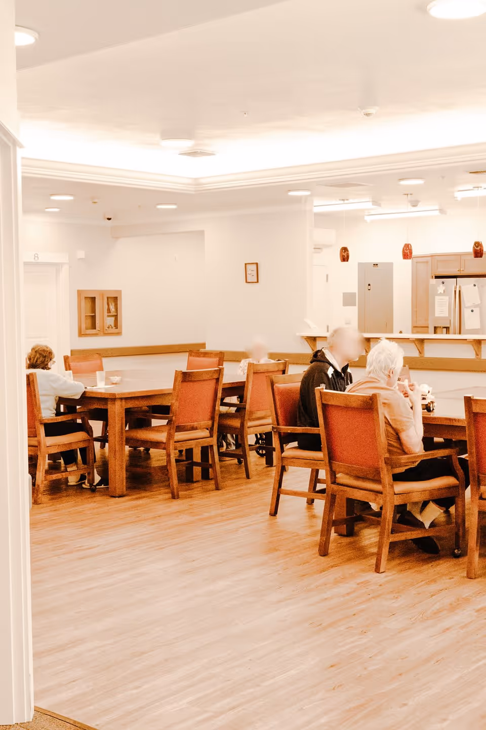 A bright communal dining room with older adults sitting at wooden tables and chairs.