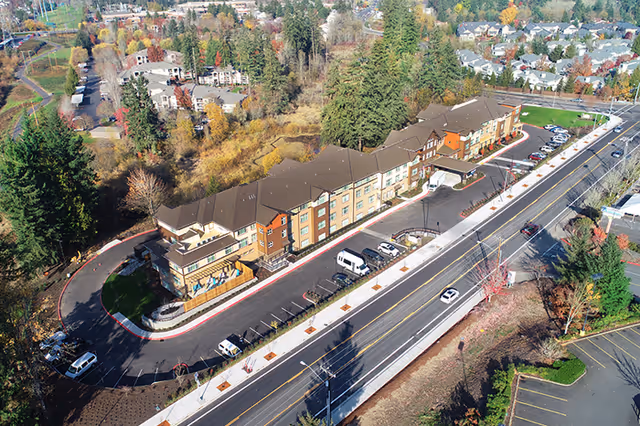Aerial view of a large multi-story senior living facility named Morning Star Of Beaverton, surrounded by trees and residential neighborhoods. The building has a brown roof and beige exterior with multiple windows. There is a curved driveway with parked vehicles in front of the building, and a main road running alongside the property.