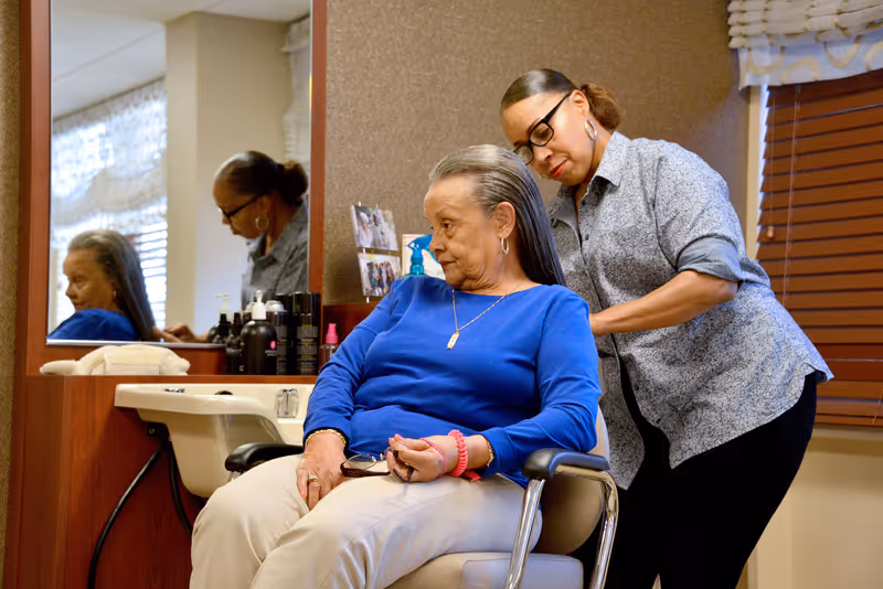An elderly woman with long gray hair sits in a salon chair wearing a blue top and beige pants, while a hairstylist standing behind her styles her hair. The setting appears to be a salon area with a large mirror, hair care products on the counter, and a window with blinds and a valance.