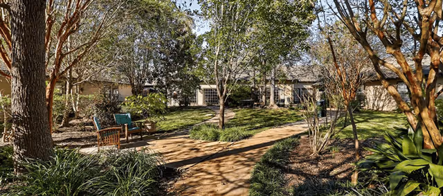 Sunlit courtyard with a winding path, trees, landscaping, a bench and a low building in the background.