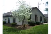 Single-story building with gray siding surrounded by green grass and bushes, featuring a small tree with white blossoms in the foreground under a clear sky.