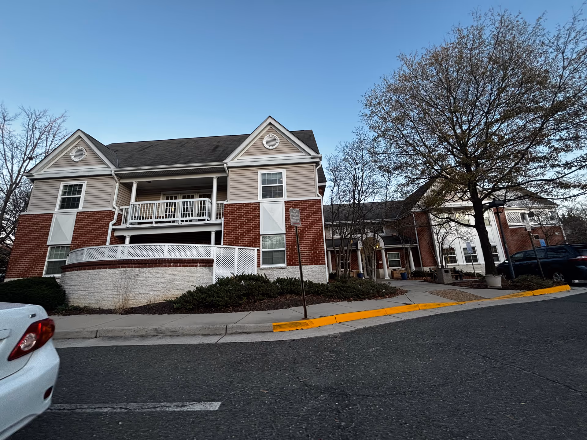 Two-story brick-and-siding senior living building with balconies, an entrance canopy, trees, and parked cars in front.