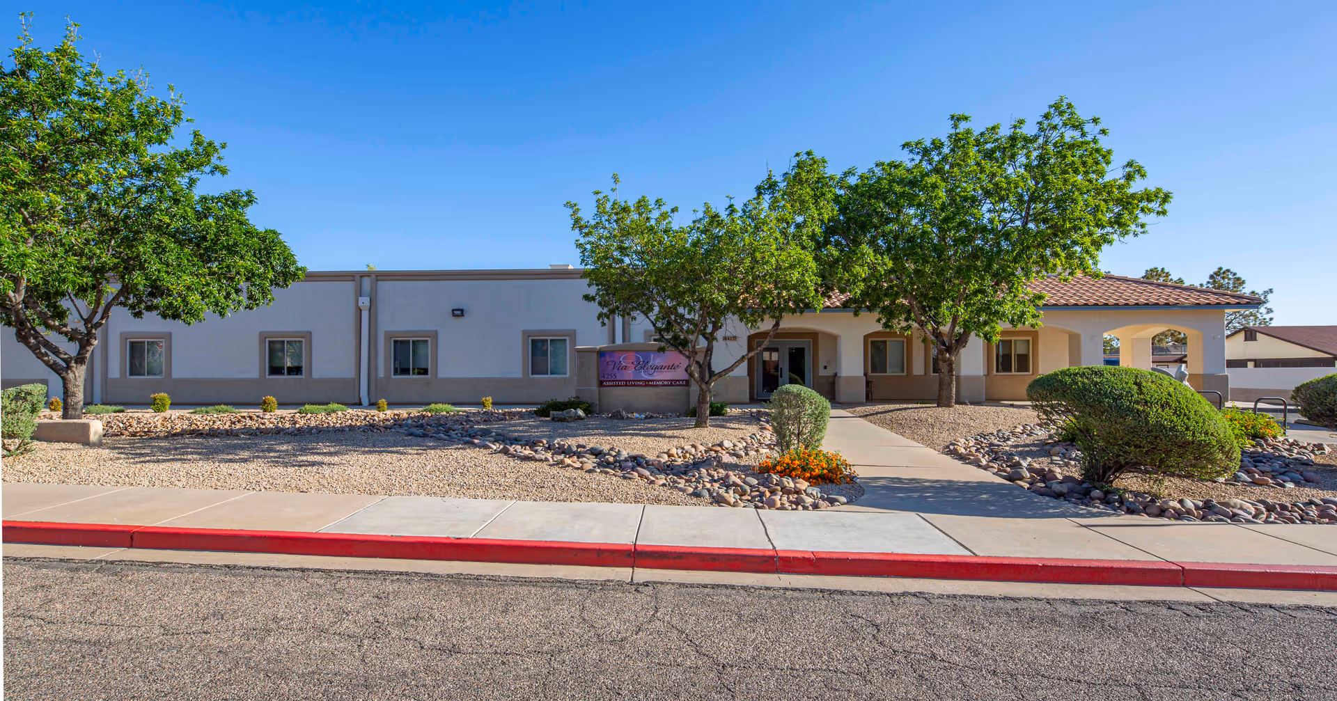Exterior view of Via Elegante Assisted Living Sierra Vista Highlands building with a clear blue sky, trees, shrubs, and a walkway leading to the entrance.