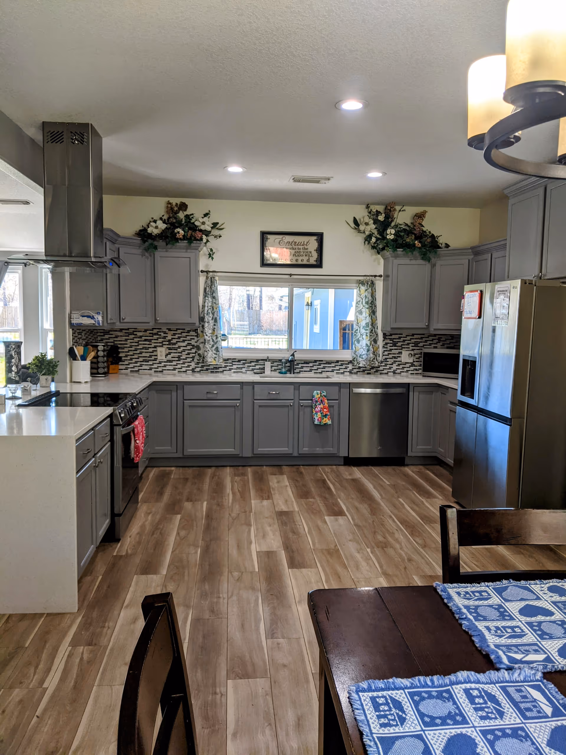A modern kitchen with gray cabinets, a stainless steel refrigerator, dishwasher, and stove. The backsplash features a mosaic tile design, and there are floral decorations above the cabinets. A window with floral curtains is centered above the sink. The floor is wood-patterned, and part of a dining table with blue and white placemats is visible in the foreground.