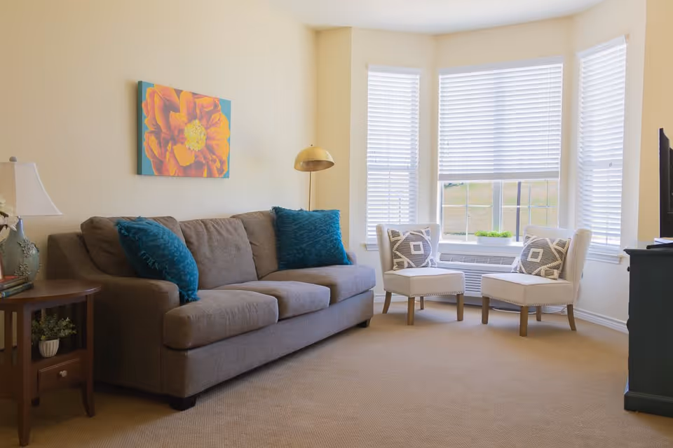 A bright living room with a gray sofa adorned with two teal pillows, two white cushioned chairs with patterned pillows, a wooden side table with a lamp and books, a floor lamp with a gold shade, and a large window with white blinds letting in natural light. A colorful flower painting hangs on the wall above the sofa.