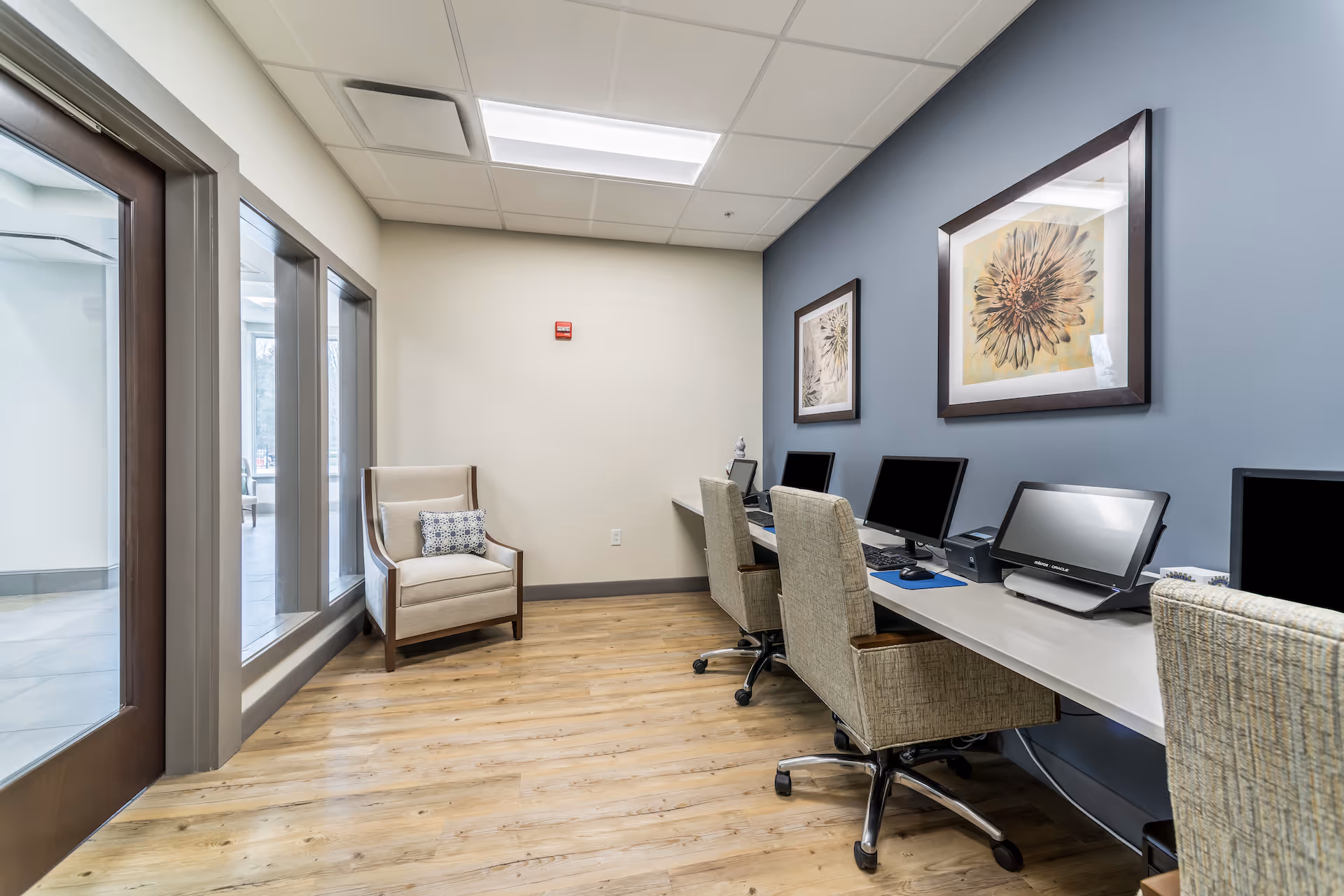 A small computer room in Gambrill Gardens Senior Apartments with three computer workstations along a blue accent wall, each with a chair on wheels. Two framed floral artworks hang above the desks. A beige armchair with a patterned pillow is placed near a set of windows and a wooden door. The room has light wood flooring and a white ceiling with fluorescent lighting.