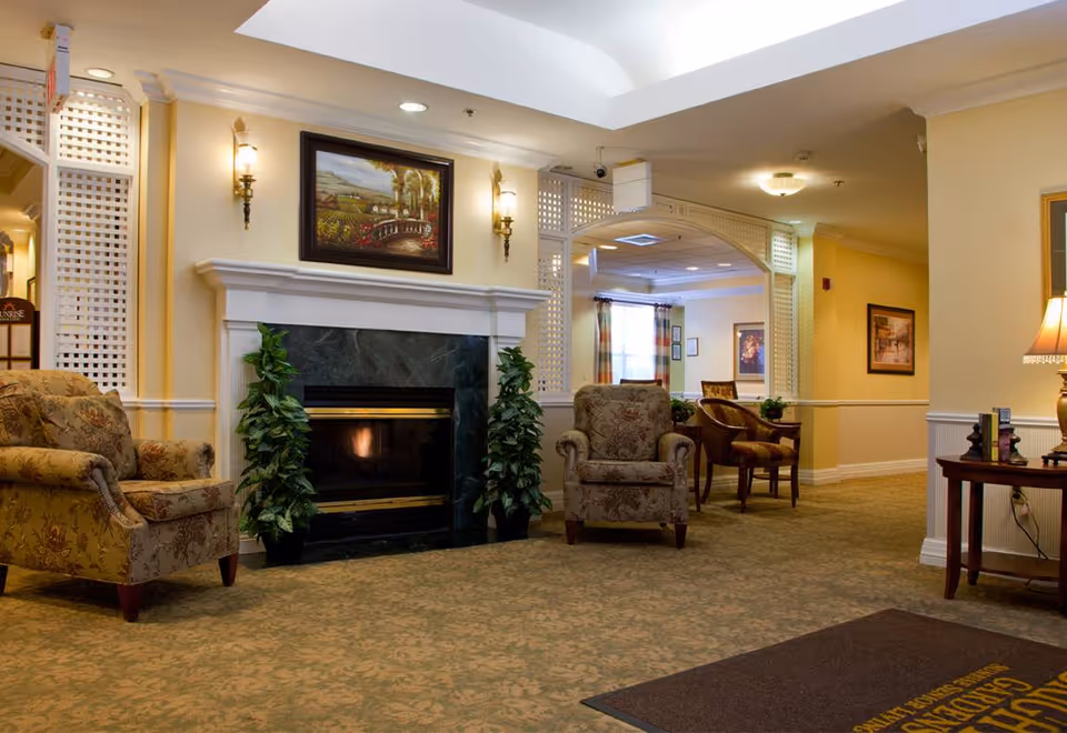A cozy senior living facility common area with two patterned armchairs facing a fireplace with a dark marble surround. Above the fireplace is a framed landscape painting and two wall sconces. The room has beige walls, carpeted floor, and decorative white lattice partitions. In the background, there is a seating area with chairs and a window with curtains.