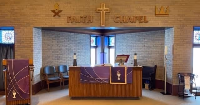 Small chapel interior with an altar draped in purple cloth, a wooden cross and the words 'FAITH CHAPEL' on a brick wall.