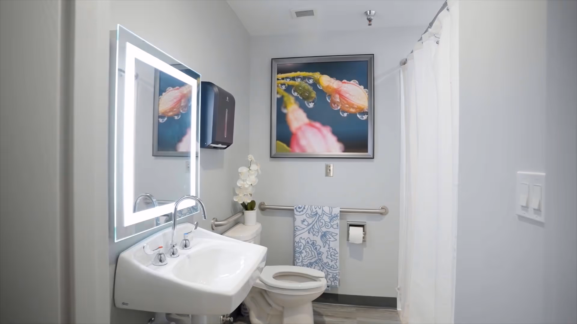A clean and modern bathroom featuring a white sink with chrome faucets, an illuminated rectangular mirror above the sink, a toilet with the seat up, a towel rack with a blue and white patterned towel, a white shower curtain, and a framed close-up photo of a flower with water droplets on the wall.