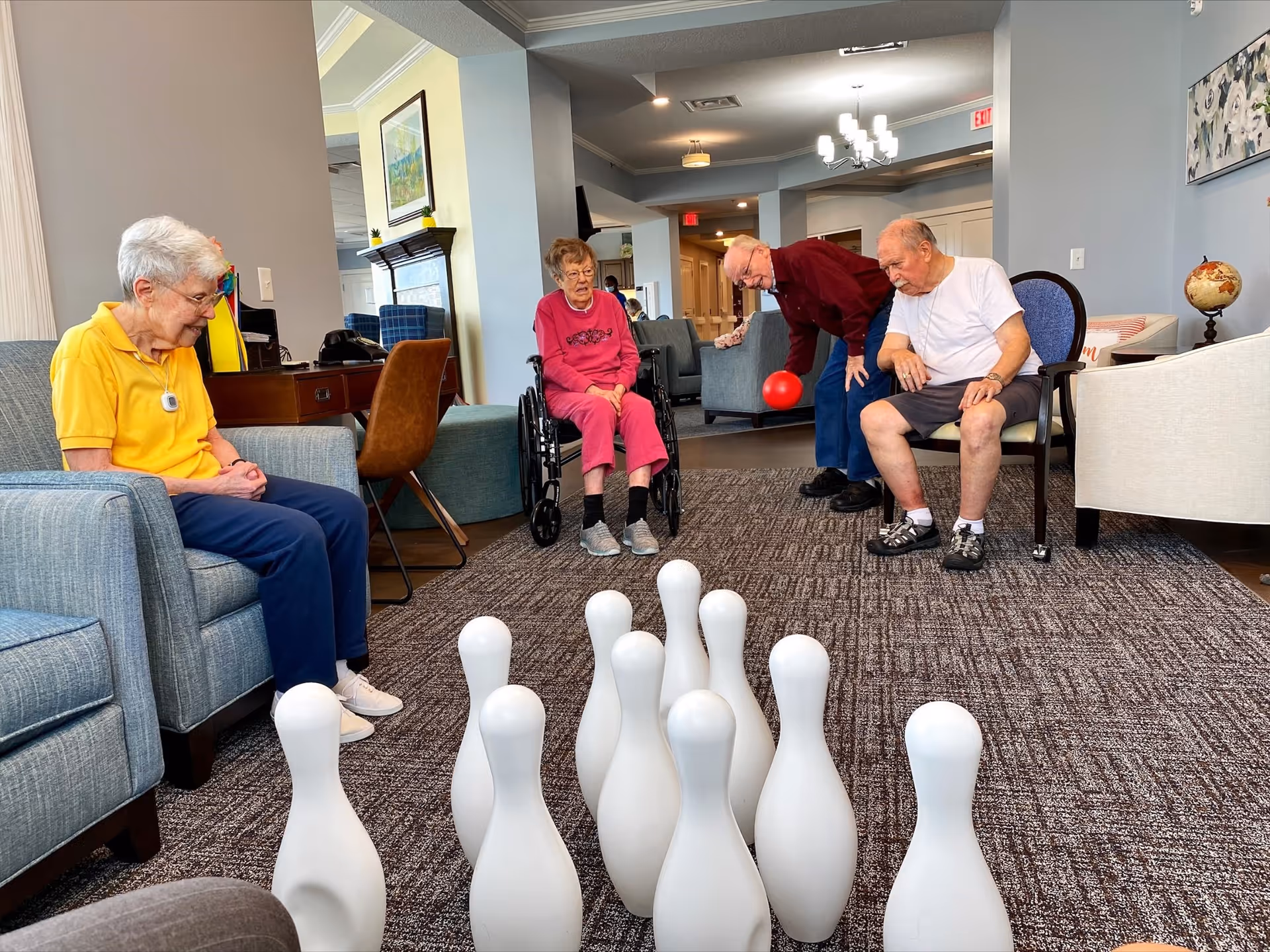 Four elderly individuals in a senior living facility common area playing a game of indoor bowling with white pins set up on the carpet. One person is in a wheelchair, another is about to roll a red ball towards the pins, and two others are seated watching the game. The room has comfortable seating, light blue walls, and a globe on a side table.