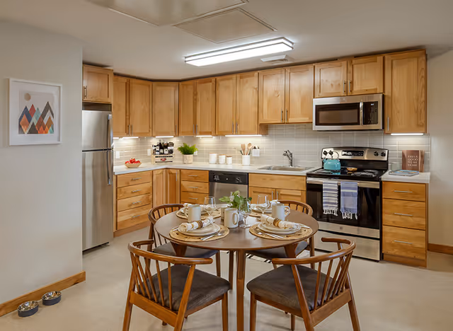 A modern kitchen with wooden cabinets, stainless steel refrigerator, stove, microwave, and dishwasher. A round dining table with four wooden chairs is set with plates, cups, and napkins. The kitchen is well-lit with under-cabinet lighting and a ceiling light. There is a framed abstract art piece on the wall and two pet bowls on the floor near the corner.