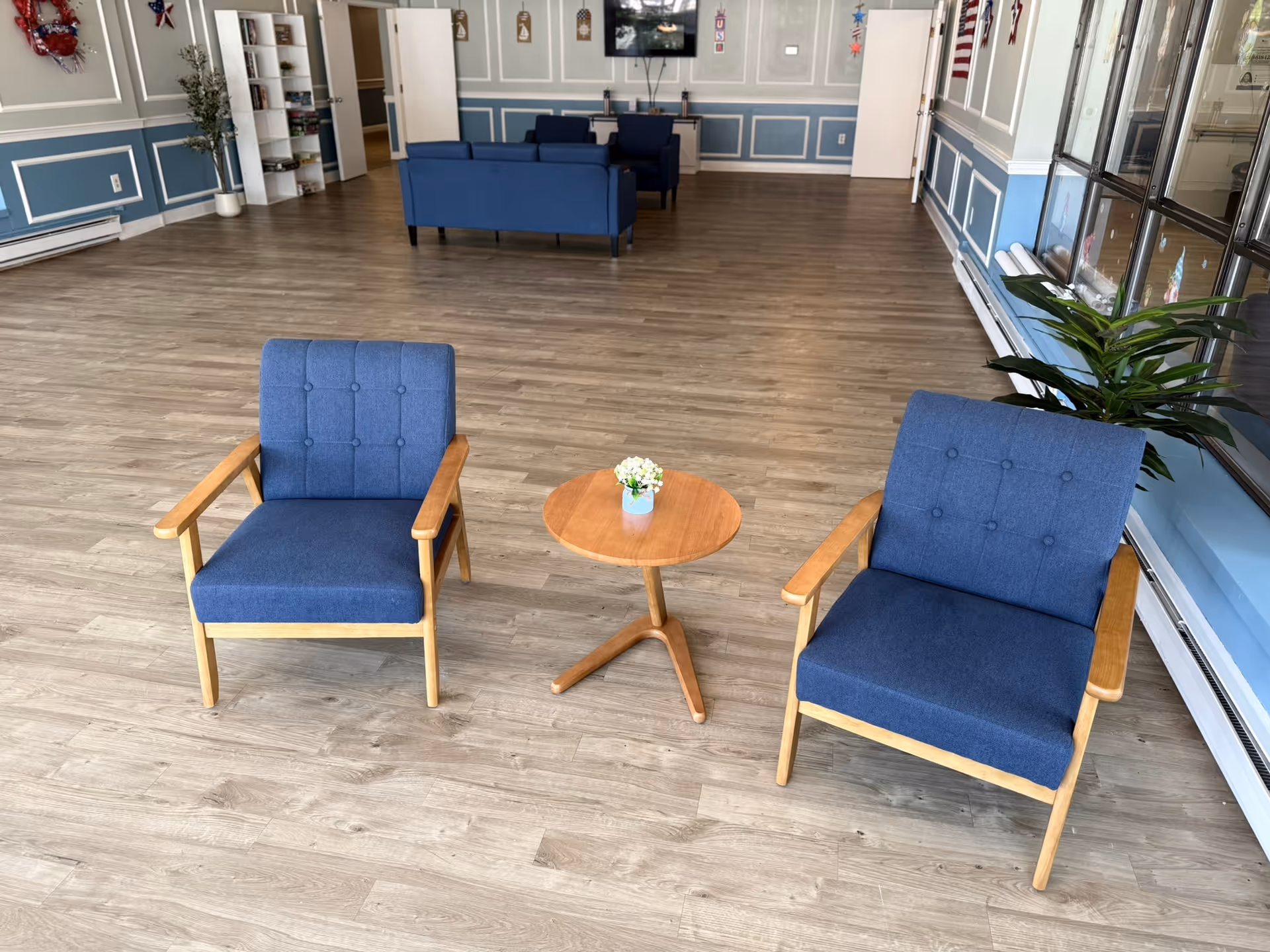 A spacious room with light wood flooring and blue walls featuring white trim. In the foreground, there are two blue cushioned wooden armchairs with a small round wooden table between them, holding a small potted plant. In the background, there are more blue sofas, a white bookshelf, and a wall-mounted TV. The room has large windows on the right side and some decorative items on the walls.