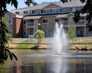 A multi-story residential building with balconies overlooking a pond with a water fountain in the center, surrounded by trees and greenery.