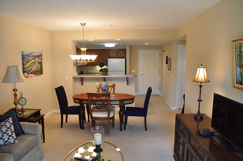 Interior view of a senior living apartment at Moravian Village of Bethlehem featuring a dining area with a round wooden table and four chairs, a kitchen with wooden cabinets and a white refrigerator in the background, a living area with a beige sofa, a side table with a lamp and clock, a TV on a wooden dresser, and warm lighting from ceiling and table lamps.
