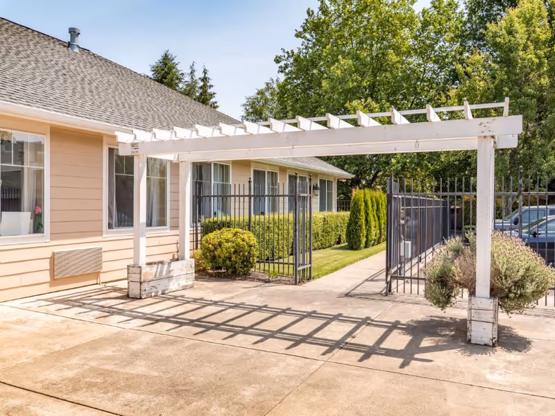 Outdoor view of a pathway next to a beige building with multiple windows. A white wooden pergola structure stands over the concrete path, casting shadows on the ground. There is a black metal fence and gate along the pathway, with green bushes and trees in the background. Several parked cars are visible beyond the fence.