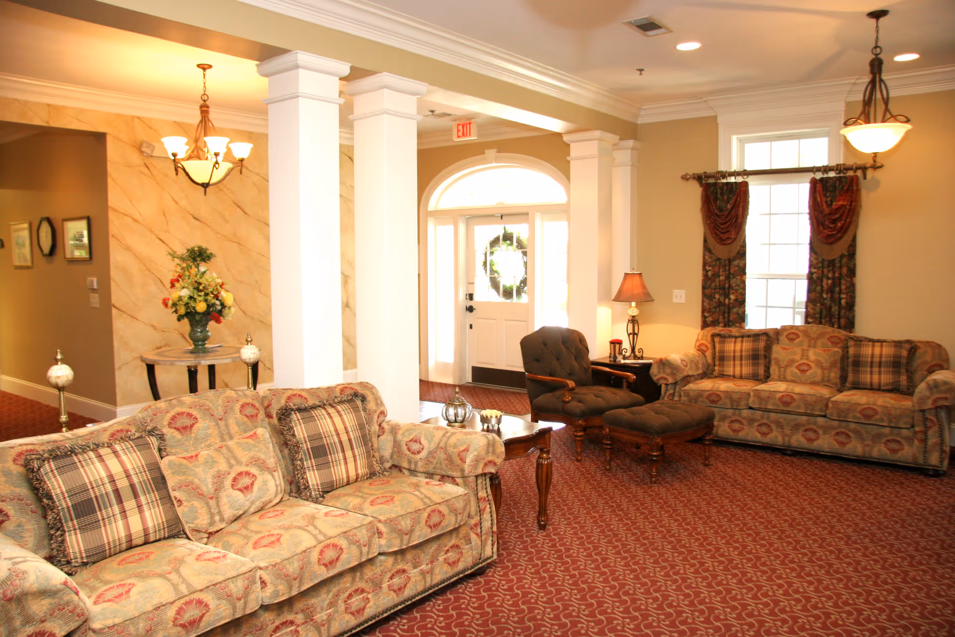 A cozy living room area with two patterned sofas, a cushioned armchair with matching ottoman, a wooden coffee table, and a side table with a lamp. The room features a red patterned carpet, beige walls, white columns, and a front door with a wreath. There are windows with floral curtains and a chandelier hanging from the ceiling.