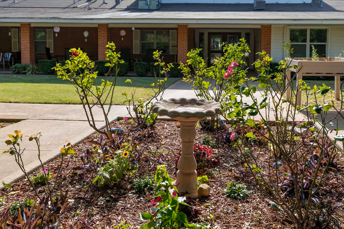 A small courtyard garden with a stone birdbath and flowering plants in front of a single-story assisted living building.