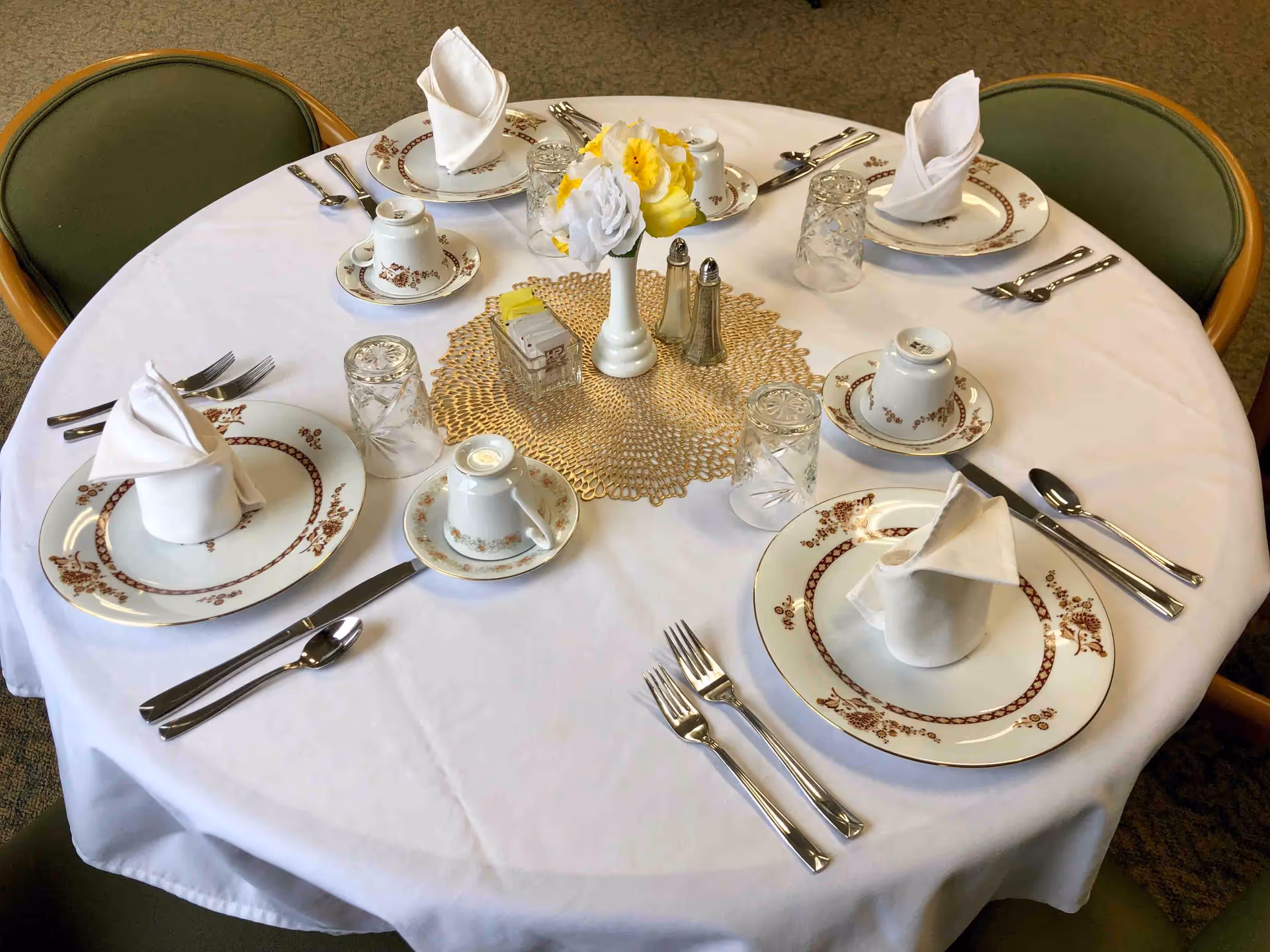 A round dining table set for five with white tablecloth, floral-patterned plates, cups, silverware, folded white napkins, glass tumblers, a small vase with yellow and white flowers, salt and pepper shakers, and a container with sugar packets.